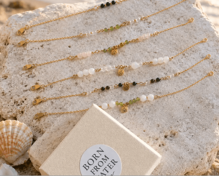 Gold-filled gemstone bracelets arranged on stone beside Born From Water packaging